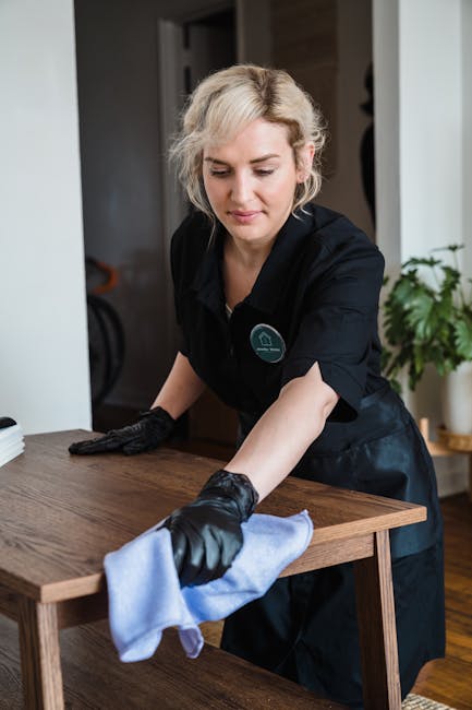 A woman wearing a black cleaning uniform with gloves is engaged in domestic surface cleaning inside a modern residential space. She is wiping a wooden dining table with a light blue cloth, ensuring the surface is dust-free and spotless. The table's surface appears polished and clean, with a smooth finish and natural wood grain. In the background, there is a white wall, a partially open door, and a green houseplant in a white pot, contributing to the bright and tidy atmosphere. The lighting is natural, casting soft shadows, emphasizing the cleanliness and shine of the wooden surface. This image highlights professional deep cleaning services provided by Islington Cleaners, focusing on surface maintenance and hygiene in a home setting, aligned with the upper street flat cleaning services in Islington.