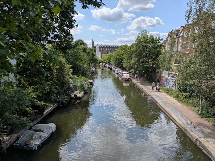 Photograph of a narrow residential canal in Islington on a bright, partly cloudy day. The canal water is calm, reflecting the blue sky and white clouds, with some ripples on the surface. On one side, there is a stone walkway lined with lush green trees and shrubs, providing shade and natural scenery. Several small boats are moored along the canal bank, some covered with protective tarps. The opposite side features a row of multi-story brick buildings with large windows, some with visible graffiti on the lower sections of walls. A few pedestrians are walking along the pathway, and parked cars are visible further down the street. Bright natural lighting highlights the vibrant greenery and the reflections in the water, creating a peaceful urban scene for the Upper Street flat cleaning services in Islington webpage by Islington Cleaners.